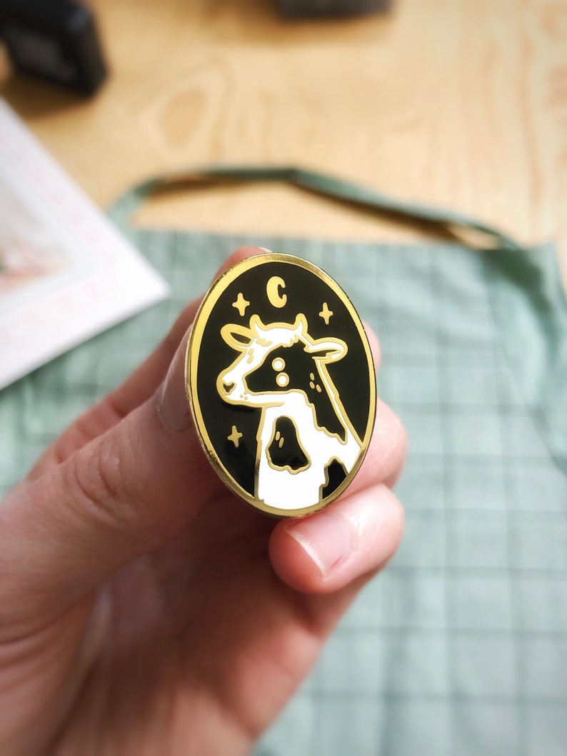 May include: A gold-colored enamel pin with a black background and a white cow with black spots. The pin has a white star on each side of the cow and the letter "C" above the cow.