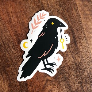 May include: A black crow with a yellow eye holds a gold key in its beak. The crow is surrounded by pink and white floral elements, stars, and a crescent moon.