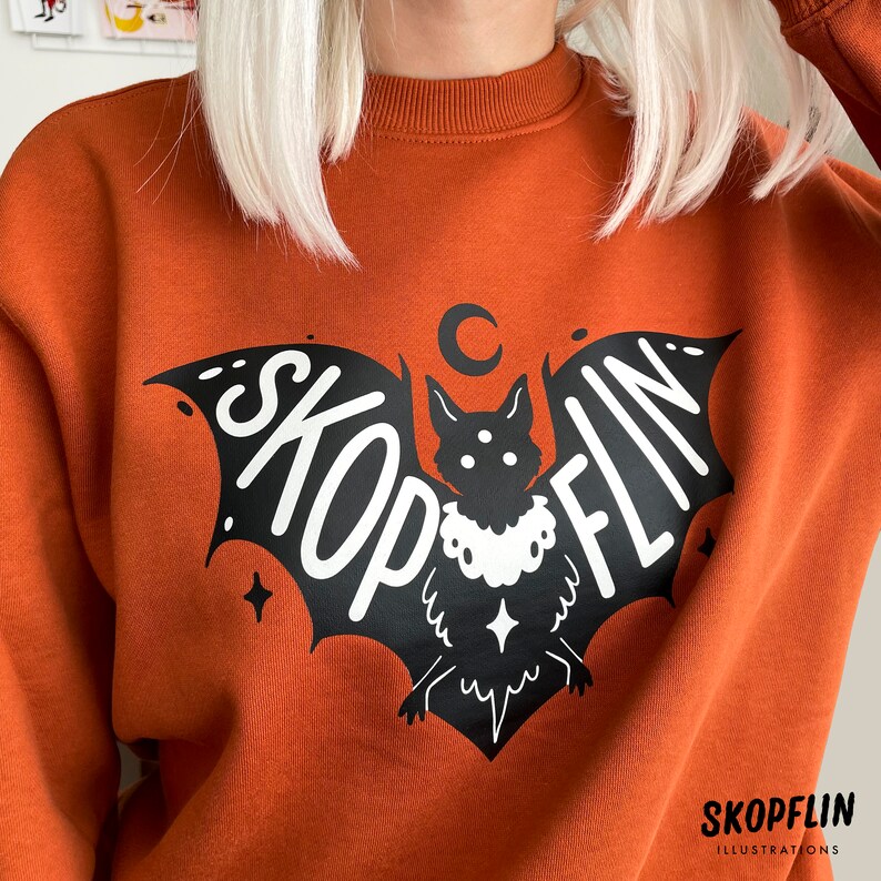 Op de afbeelding: Oranje sweatshirt met een zwarte vleermuis afbeelding. De vleermuis heeft een witte halve maan erboven en het woord "SKOPFLIN" op zijn vleugels geschreven. De tekst "SKOPFLIN ILLUSTRATIONS" is in wit gedrukt aan de onderkant van het sweatshirt.