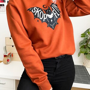 Op de afbeelding: Een oranje sweatshirt met een zwarte vleermuisprint en de tekst "SKOPFLIN" op de voorkant.