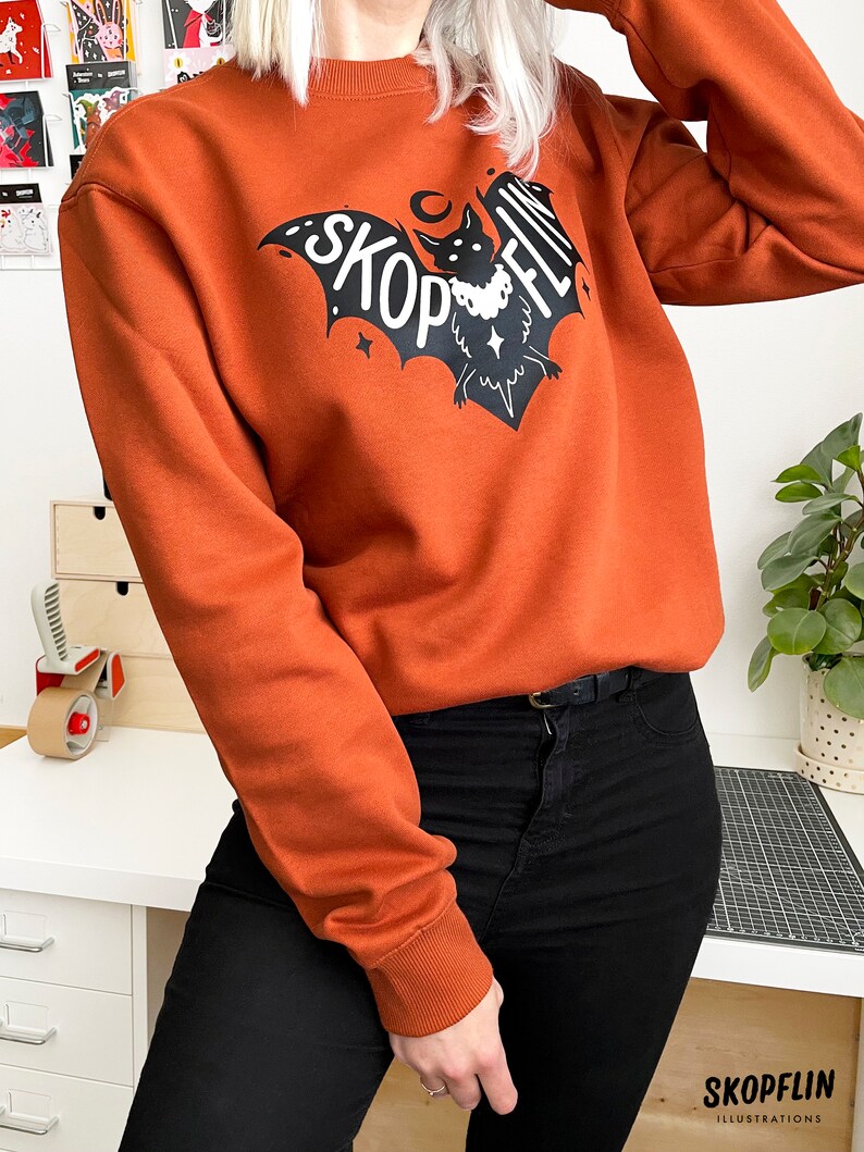 Op de afbeelding: Een oranje sweatshirt met een zwarte vleermuisprint en de tekst "SKOPFLIN" op de voorkant.