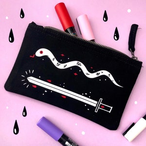 Könnte beinhalten: Schwarze Canvas-Kosmetiktasche mit einem weißen Schlangen- und Schwertmuster. Die Tasche ist mit roten, rosa und violetten Markern gefüllt.