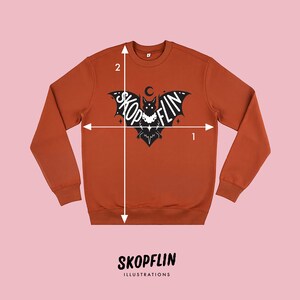 Op de afbeelding: Een roestkleurig sweatshirt met een zwarte vleermuis afbeelding. De vleermuis heeft de woorden "Skop flin" erop geschreven. Het sweatshirt wordt gemeten met twee lijnen, een horizontale en een verticale. De horizontale lijn is gemarkeerd met "1" en de verticale lijn met "2".