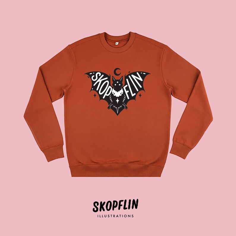 Op de afbeelding: Een roestkleurig sweatshirt met een zwarte vleermuisprint. De vleermuis heeft het woord "SKOPFLIN" op zijn vleugels staan. De vleermuis draagt een ketting met een halve maan hanger. Het sweatshirt heeft een ronde hals en lange mouwen.