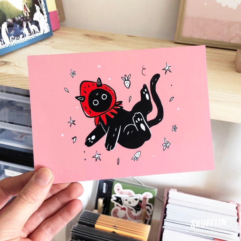 Pode incluir: Um cart&atilde;o rosa com um gato preto vestindo uma fantasia de morango vermelho. O gato est&aacute; flutuando no ar com estrelas e outros pequenos detalhes ao seu redor.