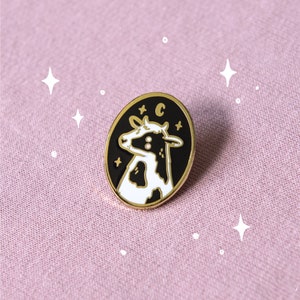 May include: Black and white enamel pin featuring a cow with a crescent moon and stars on a black background. The pin has a gold finish.
