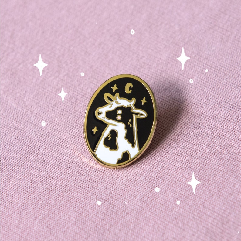 May include: Black and white enamel pin featuring a cow with a crescent moon and stars on a black background. The pin has a gold finish.