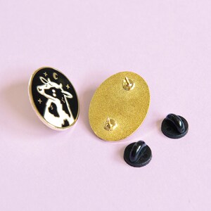 May include: A black and white enamel pin featuring a cow's head with stars and a crescent moon. The pin is on a gold background and comes with a black rubber pin back.