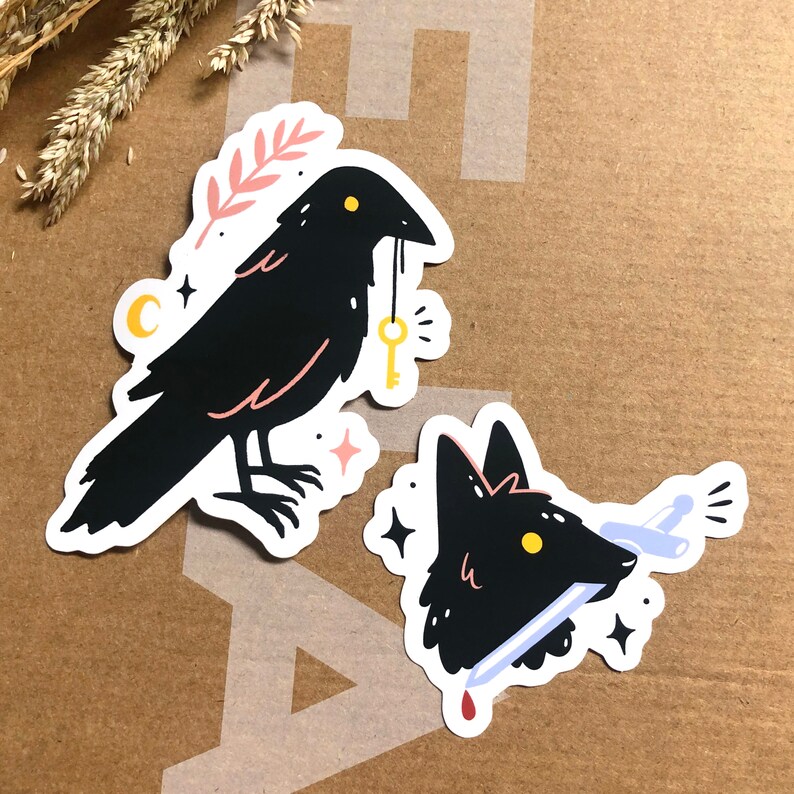 May include: Two black and white stickers featuring a crow with a key and a fox with a knife. The crow is holding a golden key in its beak and has a pink flower behind it. The fox has a red drop of blood on the knife.