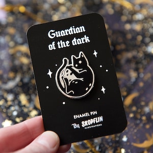 Puede incluir: Pin de esmalte en blanco y negro con un gato negro enroscado alrededor de una luna creciente con estrellas en el fondo. El pin dice "Guardian of the dark" y "Enamel Pin By Skopfelin Illustrations".
