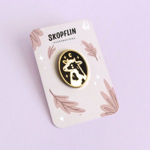 May include: A gold enamel pin featuring a black and white cow with a crescent moon and stars in a black oval. The pin is on a white card with the text "SKOPFLIN ILLUSTRATIONS" and a floral design.