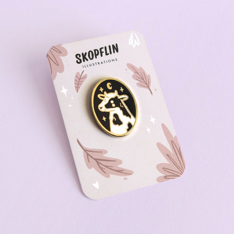 May include: A gold enamel pin featuring a black and white cow with a crescent moon and stars in a black oval. The pin is on a white card with the text "SKOPFLIN ILLUSTRATIONS" and a floral design.