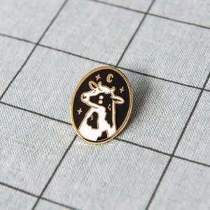 May include: A black and white enamel pin featuring a cow with stars and the letter "C" in a gold circle.