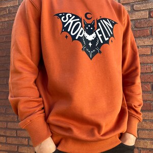 Op de afbeelding: Een oranjegekleurd sweatshirt met een zwarte vleermuis afbeelding. De vleermuis heeft een halve maan erboven en de tekst "SKOPFLIN" op zijn vleugels.