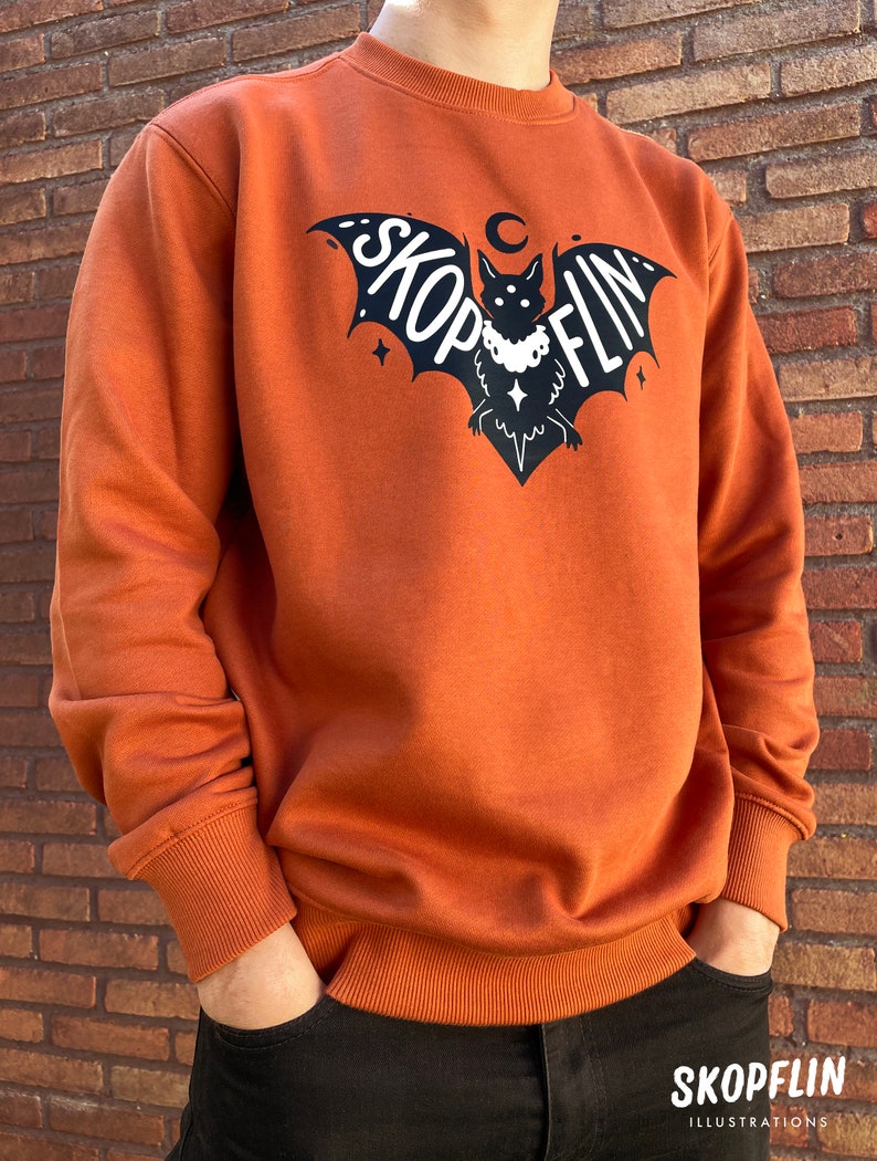Op de afbeelding: Een oranjegekleurd sweatshirt met een zwarte vleermuis afbeelding. De vleermuis heeft een halve maan erboven en de tekst "SKOPFLIN" op zijn vleugels.