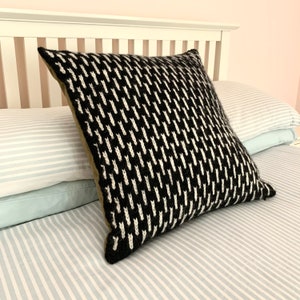 May include: A black and white patterned throw pillow with a dark green border. The pillow is on a striped bedspread.