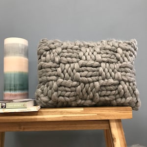May include: A large, gray, chunky knit throw pillow with a woven pattern. The pillow is sitting on a wooden bench.
