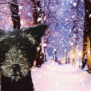 May include: A black dog's head is in the foreground, with a snowy winter forest scene in the background. The dog has a fluffy, textured appearance. The forest path is covered in snow, with falling snowflakes and streetlights.