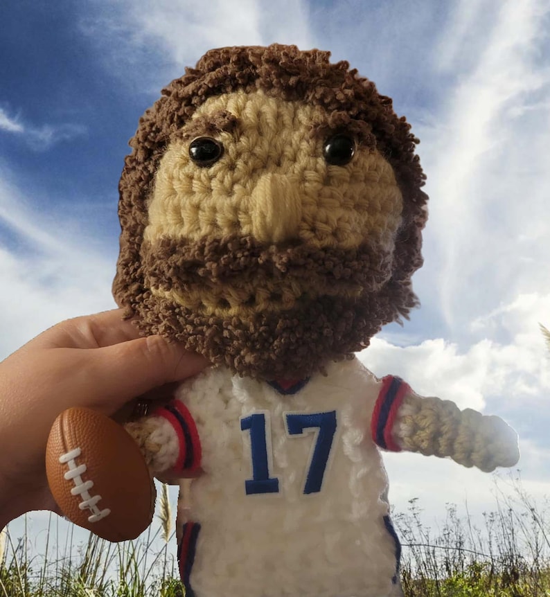 Op de afbeelding: Gehaakte pop van een American footballspeler in een wit shirt met het nummer 17 in blauw. De pop heeft een bruine baard en haar en houdt een bruine football vast. De pop staat tegen een achtergrond van een blauwe lucht.
