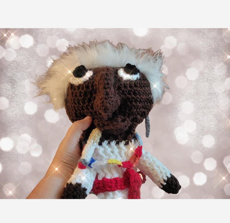 Pode incluir: Boneca de croch&ecirc; feita &agrave; m&atilde;o com rosto castanho, cabelo branco e olhos pretos. A boneca usa uma roupa branca e vermelha com detalhes coloridos. A boneca est&aacute; sendo segurada por uma m&atilde;o.