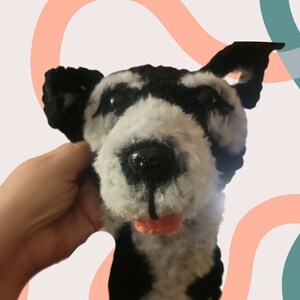 May include: A handmade plush dog head with black and white fur, black ears, and a pink tongue. The dog's eyes are black and shiny. The plushie is held by a hand.