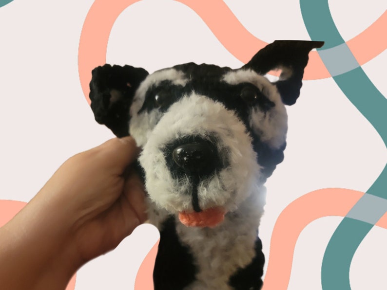 May include: A handmade plush dog head with black and white fur, black ears, and a pink tongue. The dog's eyes are black and shiny. The plushie is held by a hand.