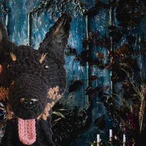 May include: A handmade, black and tan crocheted dog head with a pink tongue. The dog's face is detailed with dark eyes and a shiny black nose. The background features a textured blue wall with dark foliage and candle holders.