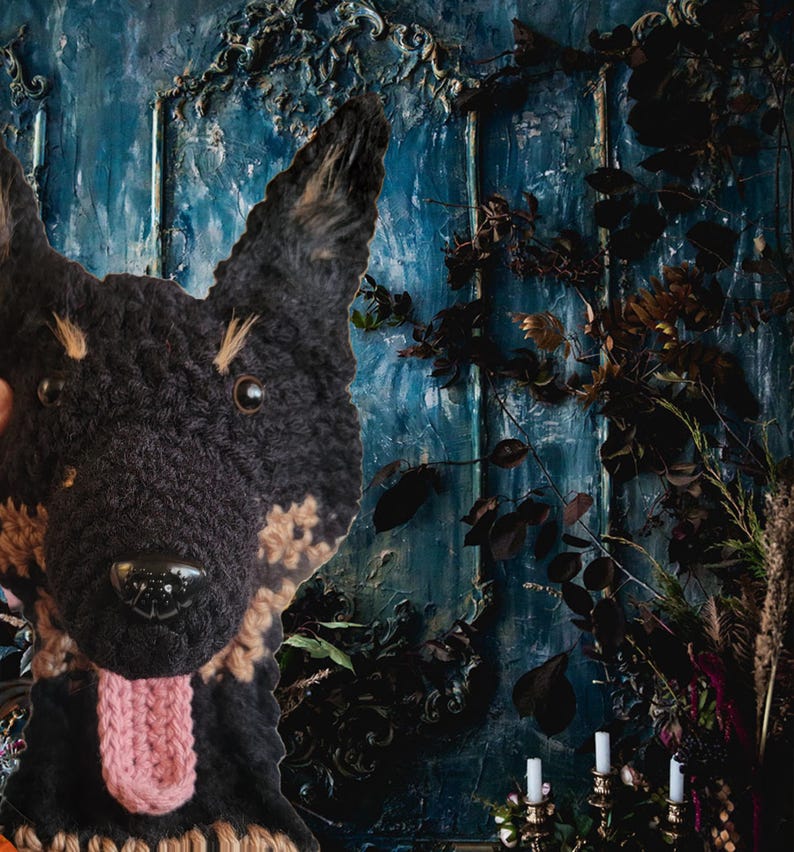 May include: A handmade, black and tan crocheted dog head with a pink tongue. The dog's face is detailed with dark eyes and a shiny black nose. The background features a textured blue wall with dark foliage and candle holders.