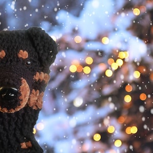 May include: A black and brown crocheted dog hat with a black nose and brown accents. The hat is designed to resemble a Rottweiler. The background features a blurred image of a Christmas tree with bokeh lights and falling snow.