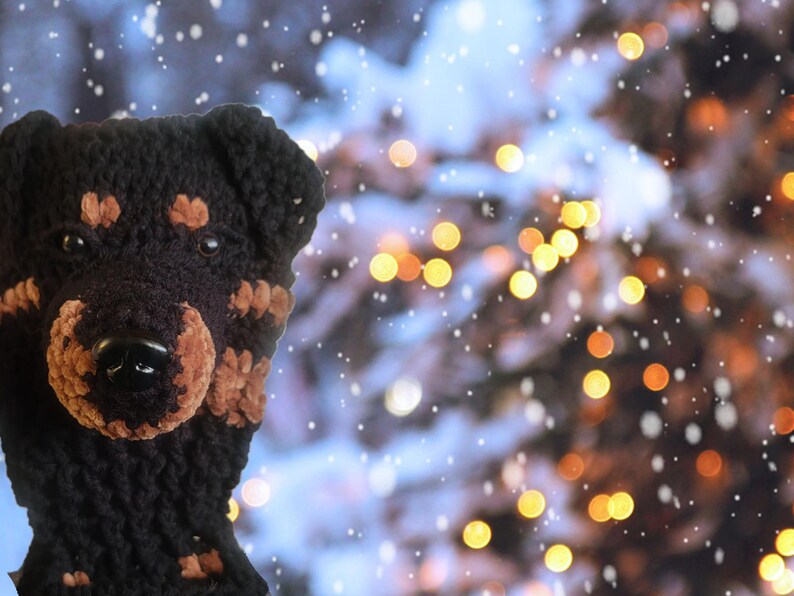 May include: A black and brown crocheted dog hat with a black nose and brown accents. The hat is designed to resemble a Rottweiler. The background features a blurred image of a Christmas tree with bokeh lights and falling snow.