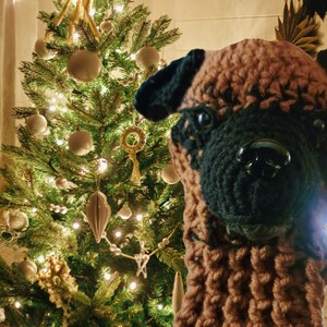 May include: A handcrafted, brown and black crocheted dog head with a black nose and eyes. The dog head is positioned in front of a decorated Christmas tree with white ornaments and lights, creating a festive holiday scene.