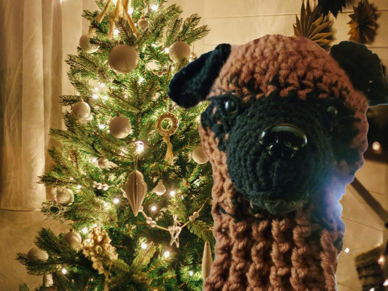 May include: A handcrafted, brown and black crocheted dog head with a black nose and eyes. The dog head is positioned in front of a decorated Christmas tree with white ornaments and lights, creating a festive holiday scene.