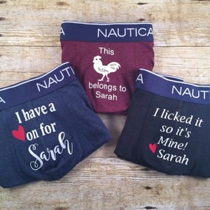 May include: Three pairs of Nautica boxer briefs in navy, burgundy, and black. Each pair has a white graphic and text. The navy pair says "I have a on for Sarah" with a red heart. The burgundy pair says "This belongs to Sarah". The black pair says "I licked it so it's Mine! Sarah".