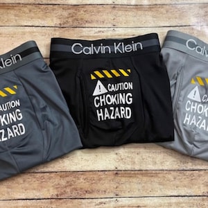 May include: Three pairs of Calvin Klein boxer briefs in gray, black, and gray. Each pair has the text "CAUTION CHOKING HAZARD" with a warning symbol. The waistband features the Calvin Klein logo.