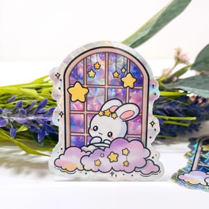 Puede incluir: Una pegatina holográfica con un conejito de dibujos animados mirando por una ventana. La ventana tiene un fondo morado y plateado con estrellas colgantes. El conejito es blanco con una corona de estrellas y está sentado en una nube con estrellas.