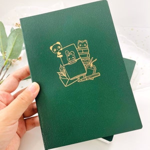 Puede incluir: Un diario verde oscuro con un diseño dorado en relieve que presenta un oso, un conejito, un gato, libros y artículos de papelería. La palabra "ECHO" es visible en el diario. El diario se sostiene en una mano.