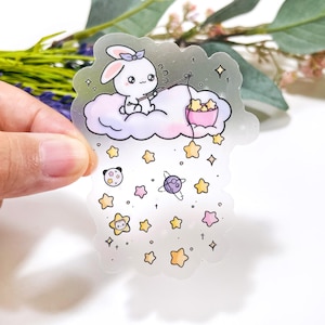 May include: A clear sticker featuring a cartoon bunny on a cloud, fishing for stars. The bunny wears a bow and sits on a fluffy cloud. Stars and planets are scattered around the bunny, with a pink bowl catching the stars.