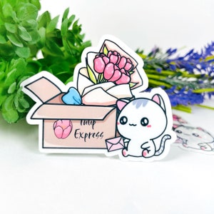 Puede incluir: Un gato blanco con orejas grises sostiene un sobre rosa frente a una caja con un ramo de tulipanes rosas dentro. La caja está etiquetada con "Tulip Express".
