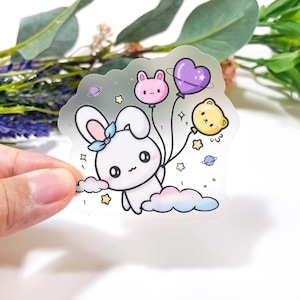 May include: A clear sticker featuring a white cartoon bunny holding three balloons: a pink bunny, a purple heart, and a yellow bear. The bunny is on a cloud, with stars and planets around it. The bunny has a blue bow.