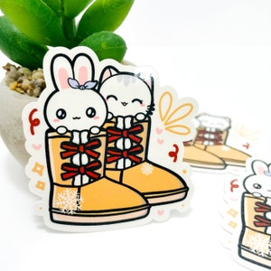 May include: A sticker featuring a white bunny and a white cat wearing yellow boots with red bows. The boots have snowflakes on them. The sticker has a white background with a yellow, orange, and pink design.