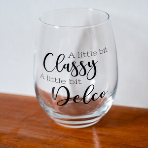 Puede incluir: Una copa de vino de vidrio transparente con el texto "A little bit Classy A little bit Delco" impreso en ella.