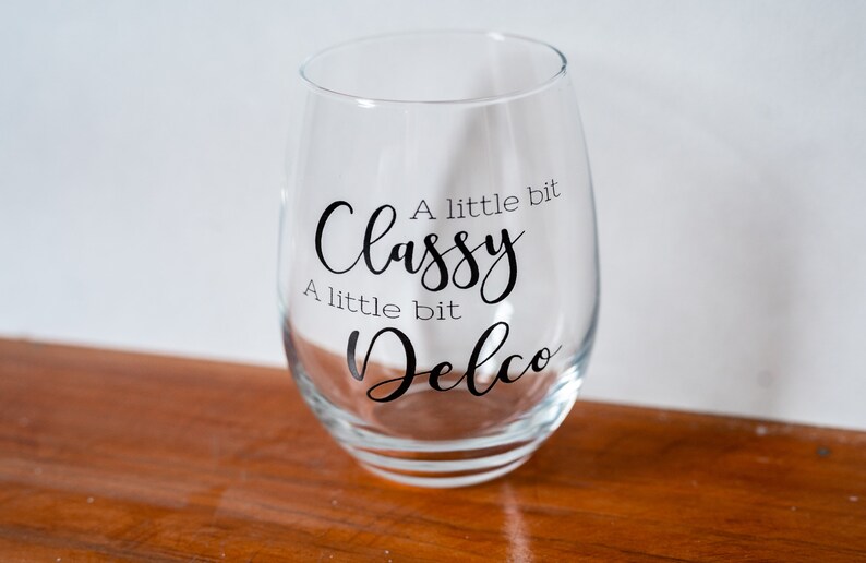 Puede incluir: Una copa de vino de vidrio transparente con el texto "A little bit Classy A little bit Delco" impreso en ella.