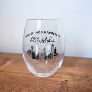 Puede incluir: Una copa de vino de vidrio transparente con un gráfico negro del horizonte de Filadelfia y el texto "Bad Things Happen in Philadelphia".