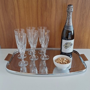 May include: A rectangular silver-toned serving tray with rounded corners and wooden handles, holding four clear crystal champagne flutes with vertical cut patterns, a bottle of sparkling wine with a floral label, and a small white bowl of nuts.