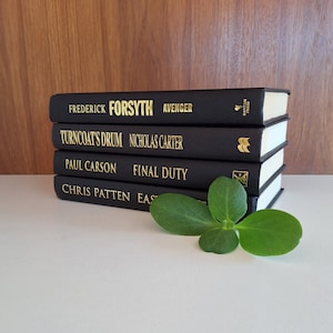 Puede incluir: Una pila de cinco libros de tapa dura negros con letras doradas en los lomos. Los t&iacute;tulos son "Avenger" de Frederick Forsyth, "Turncoat's Drum" de Nicholas Carter, "Final Duty" de Paul Carson y "East" de Chris Patten. Los libros est&aacute;n sobre una superficie blanca con una planta verde en primer plano.
