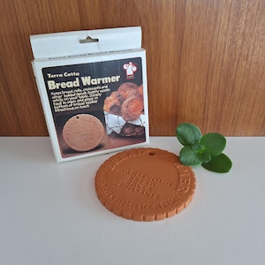 May include: A terra cotta bread warmer with a round shape and a hole for hanging. The packaging reads "Bread Warmer" and includes instructions. The warmer is designed to keep bread, rolls, and croissants warm. A small green plant is next to the warmer.
