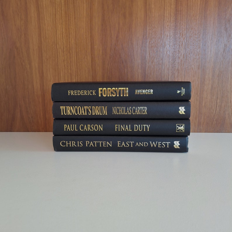 Puede incluir: Una pila de cuatro libros de tapa dura negros con letras doradas en los lomos. Los t&iacute;tulos son "Avenger" de Frederick Forsyth, "Turncoat's Drum" de Nicholas Carter, "Final Duty" de Paul Carson y "East and West" de Chris Patten.