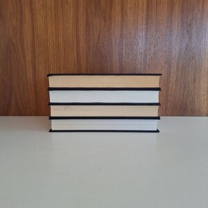 Puede incluir: Una pila de cuatro libros con cubiertas negras y p&aacute;ginas blancas. Los libros est&aacute;n dispuestos en una pila vertical sobre una superficie blanca.