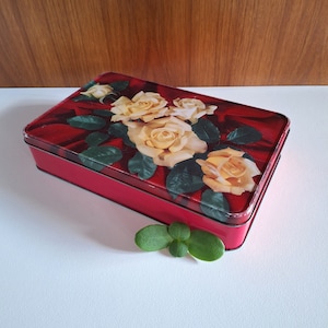 May include: Red rectangular tin box with a floral design featuring yellow roses and green leaves on a red background.