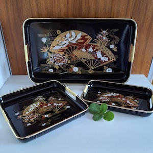 May include: A set of three black serving trays with gold trim. The trays feature a Japanese-inspired design with cranes, fans, and floral motifs in gold, red, and white. The largest tray has handles.